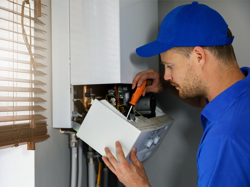 A man in a blue shirt and hat performing routine maintenance on a gas boiler.