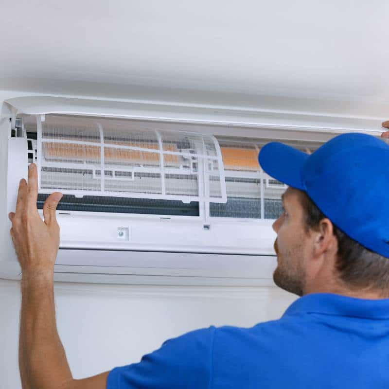 A man in a blue shirt and hat inspecting an air conditioner as part of routine HVAC maintenance.