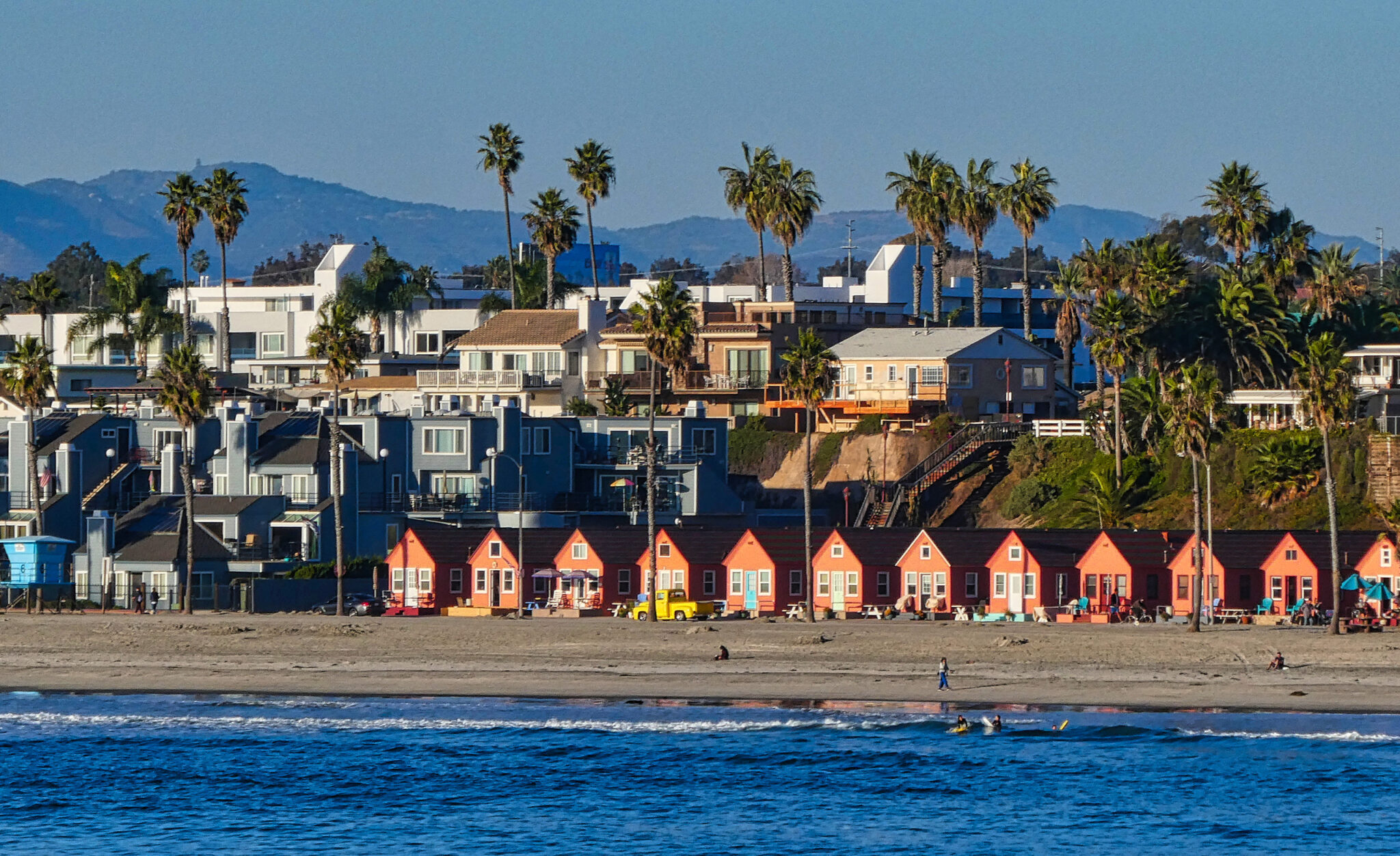 A scenic beach in Oceanside, CA, featuring houses and palm trees lining the shore under a clear blue sky.