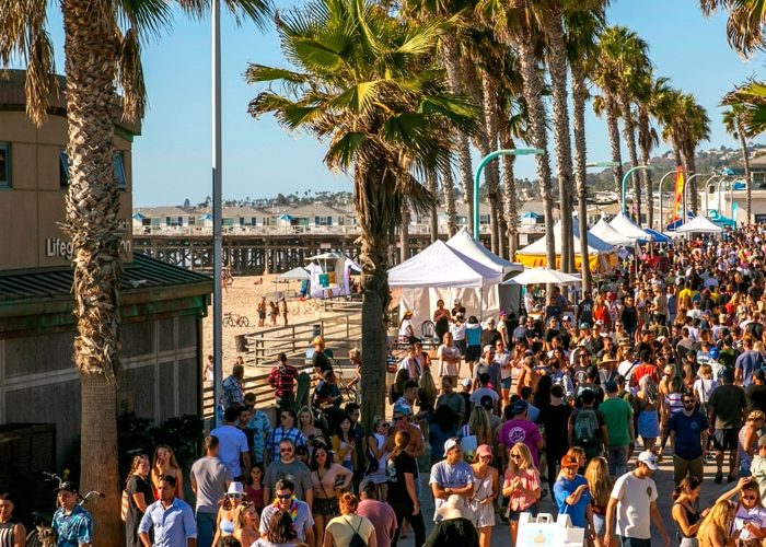 A crowd of people walking along the beach near palm trees at Pacific Beachfest in San Diego
